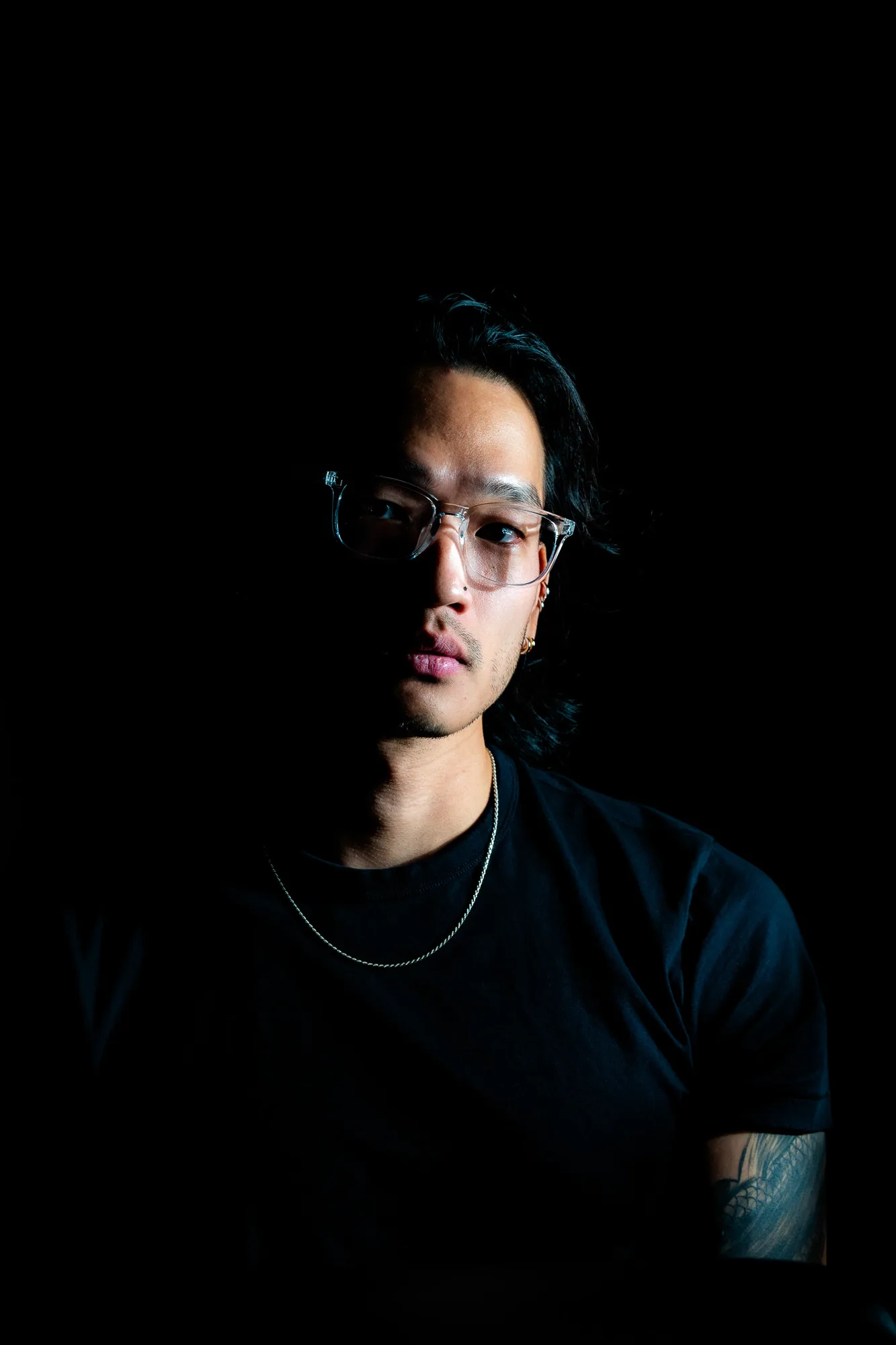 David Koi in a black t-shirt and clear-framed glasses, lit from one side against a black background.