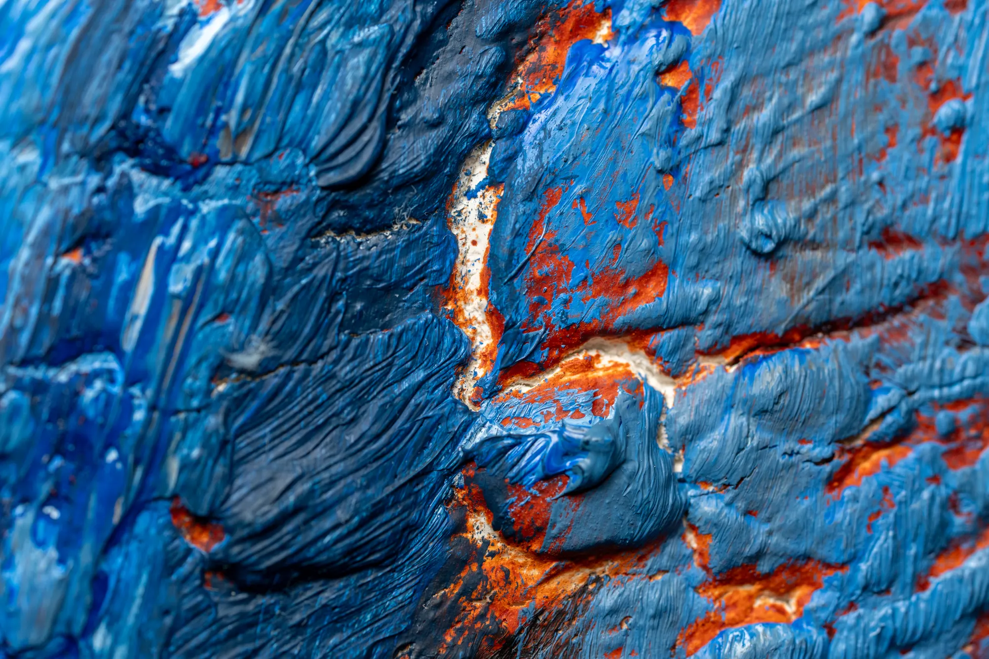 Close view of blue strokes over a red-orange underlayer that opens through the cracks.