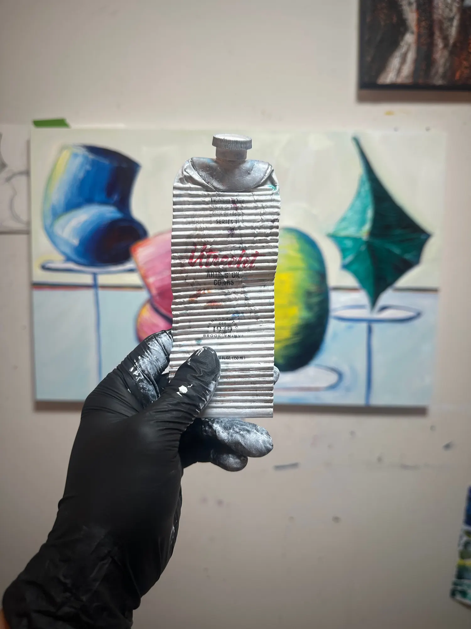 A gloved hand holding a completely flattened 150ml tube of titanium white oil paint in front of a Display Room study.
