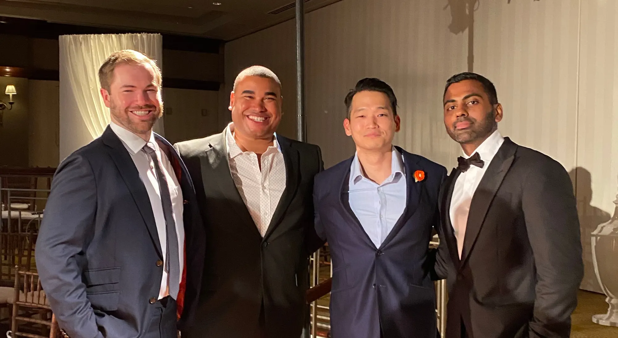 Four men in dark suits standing shoulder to shoulder at an indoor wedding reception, one wearing an orange boutonniere.