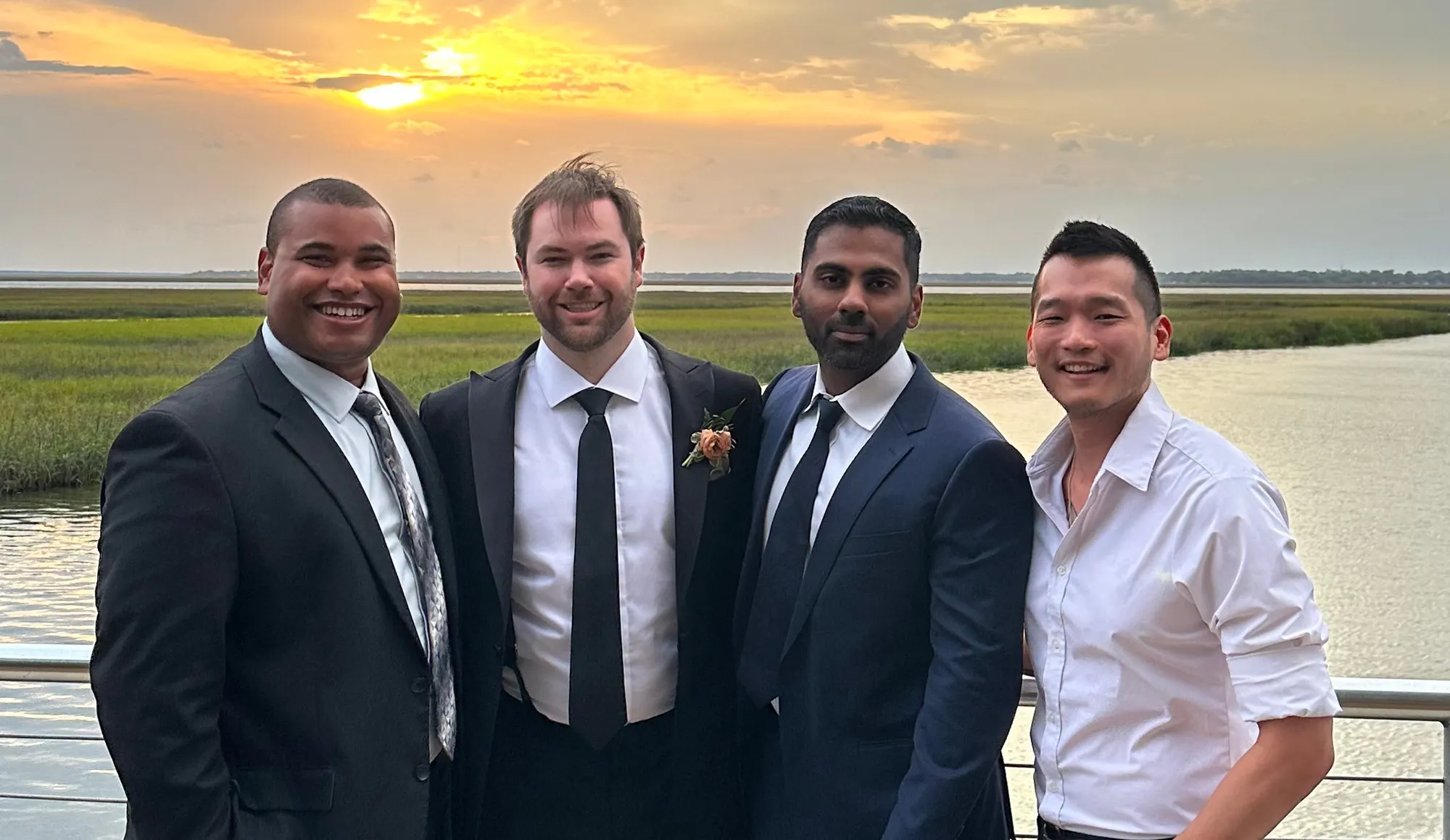Four men in dark suits on a deck at sunset, marsh grass and open water behind them.