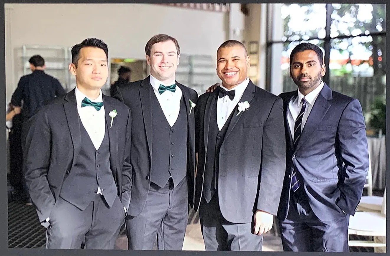 Four men in dark three-piece suits and green bow ties standing together indoors before a wedding.